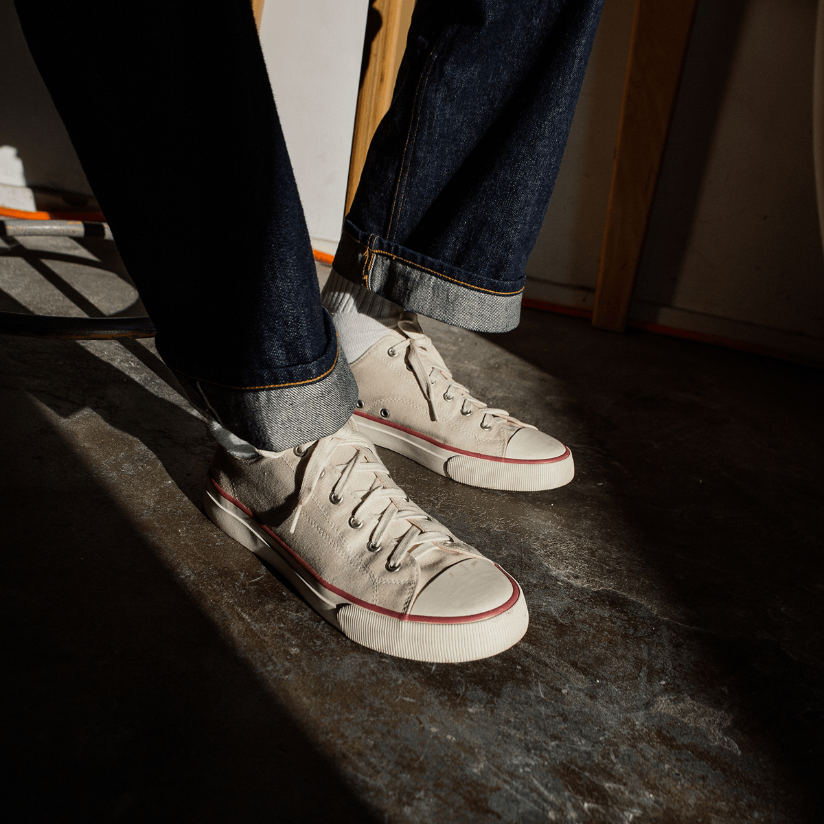 Person wearing white low-top sneakers with crimson accents and blue jeans sitting on a concrete floor.