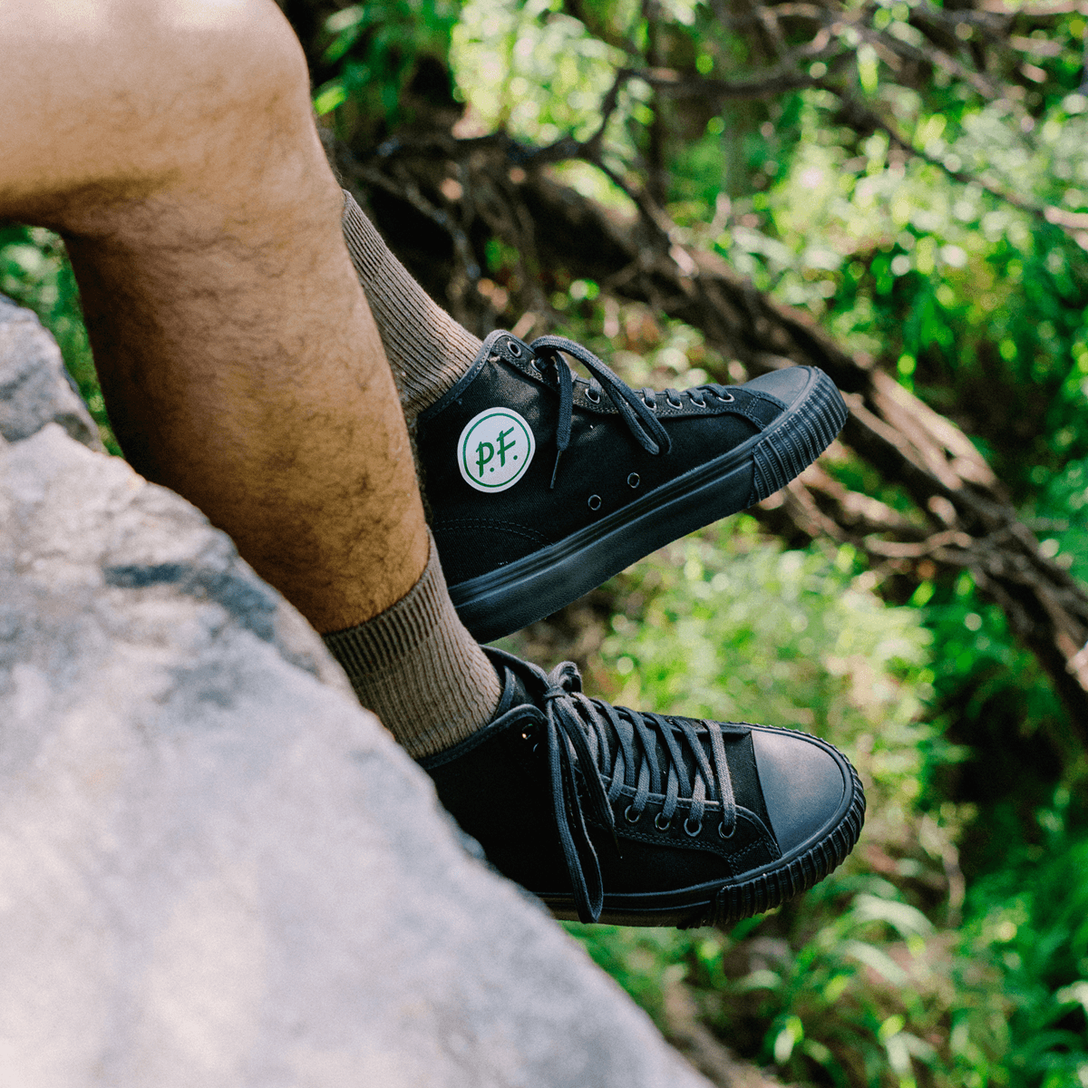 Person wearing black high-top sneakers sitting on a rock outdoors.