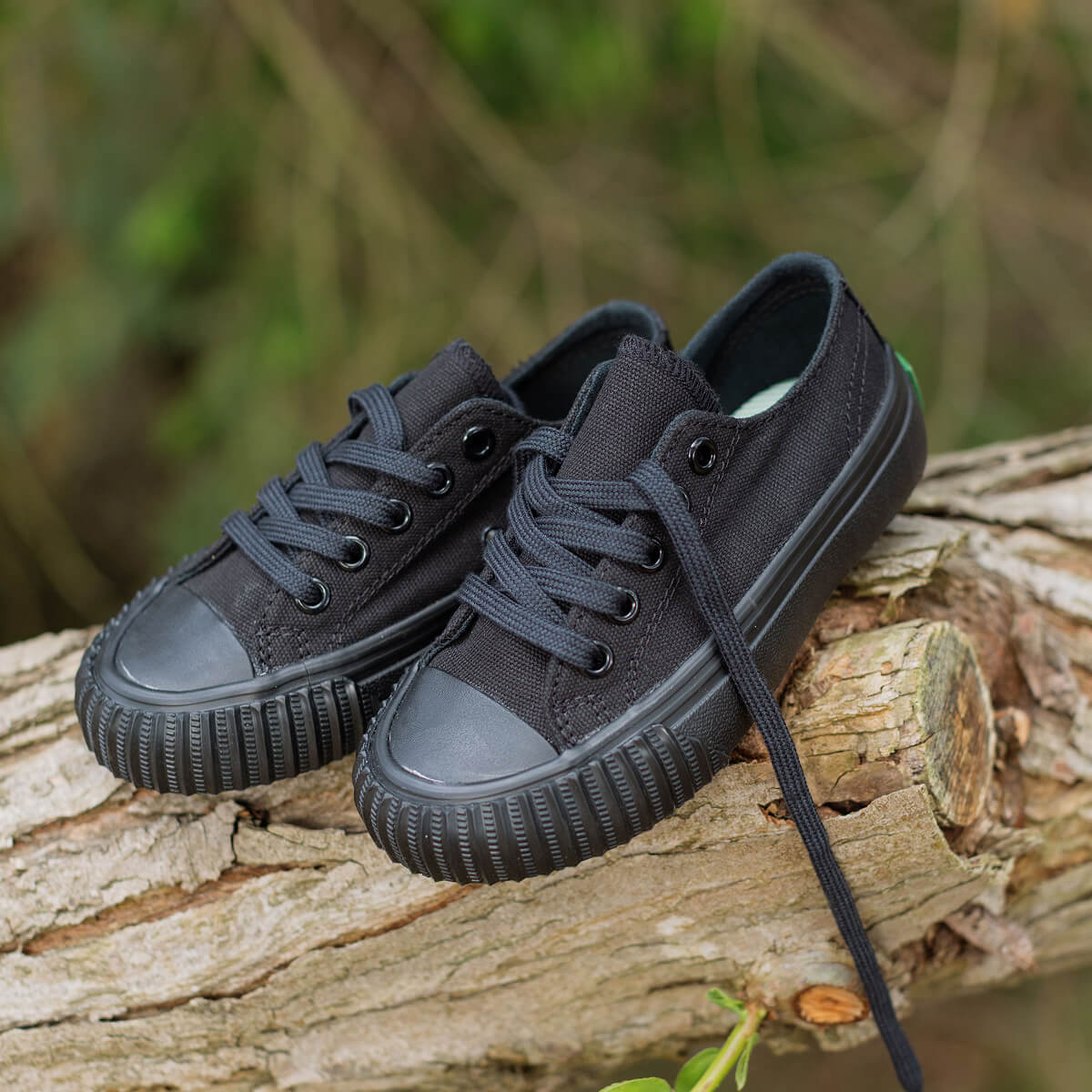 A pair of kids' black low-top sneakers resting on a weathered log with a blurred natural background.