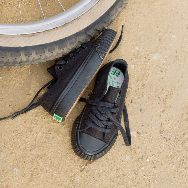 A pair of kids' black low-top sneakers with textured soles, placed on sandy ground next to a bicycle tire.