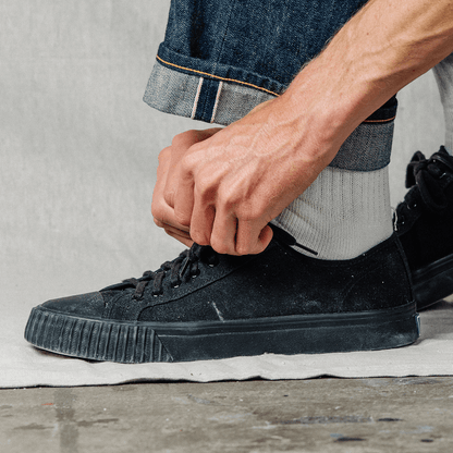 Person tying the laces of a black low-top sneaker on a neutral background.