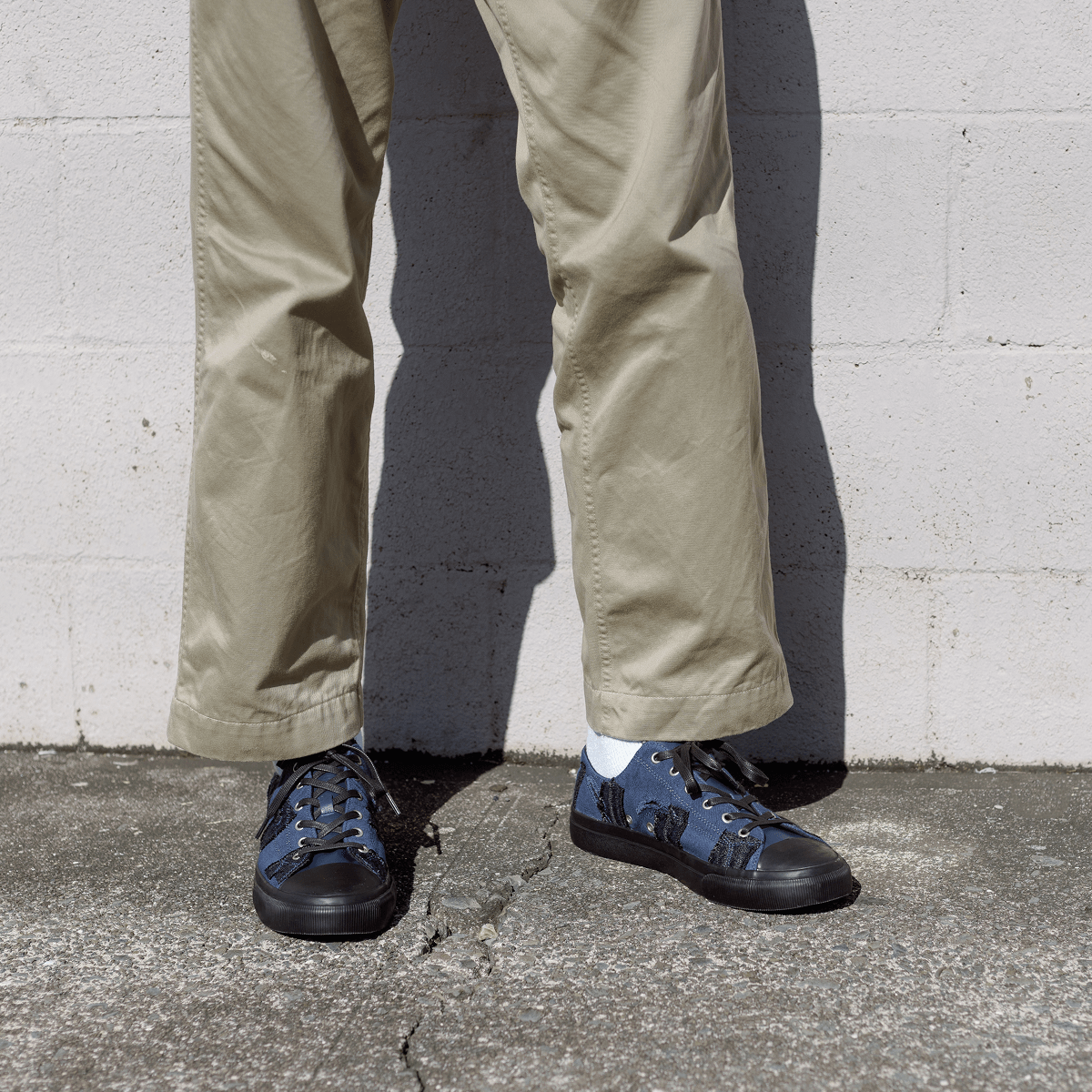 Person wearing beige pants and blue sneakers standing on a concrete surface with a white brick wall in the background.