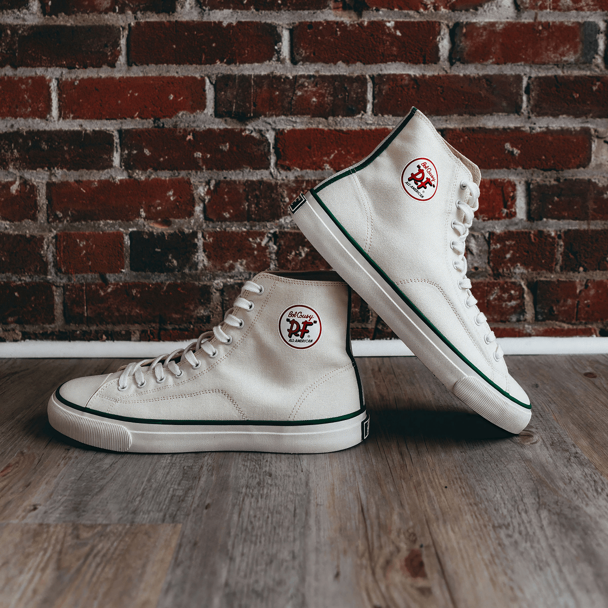White high-top sneakers with green accents on a wooden floor with a brick wall background.