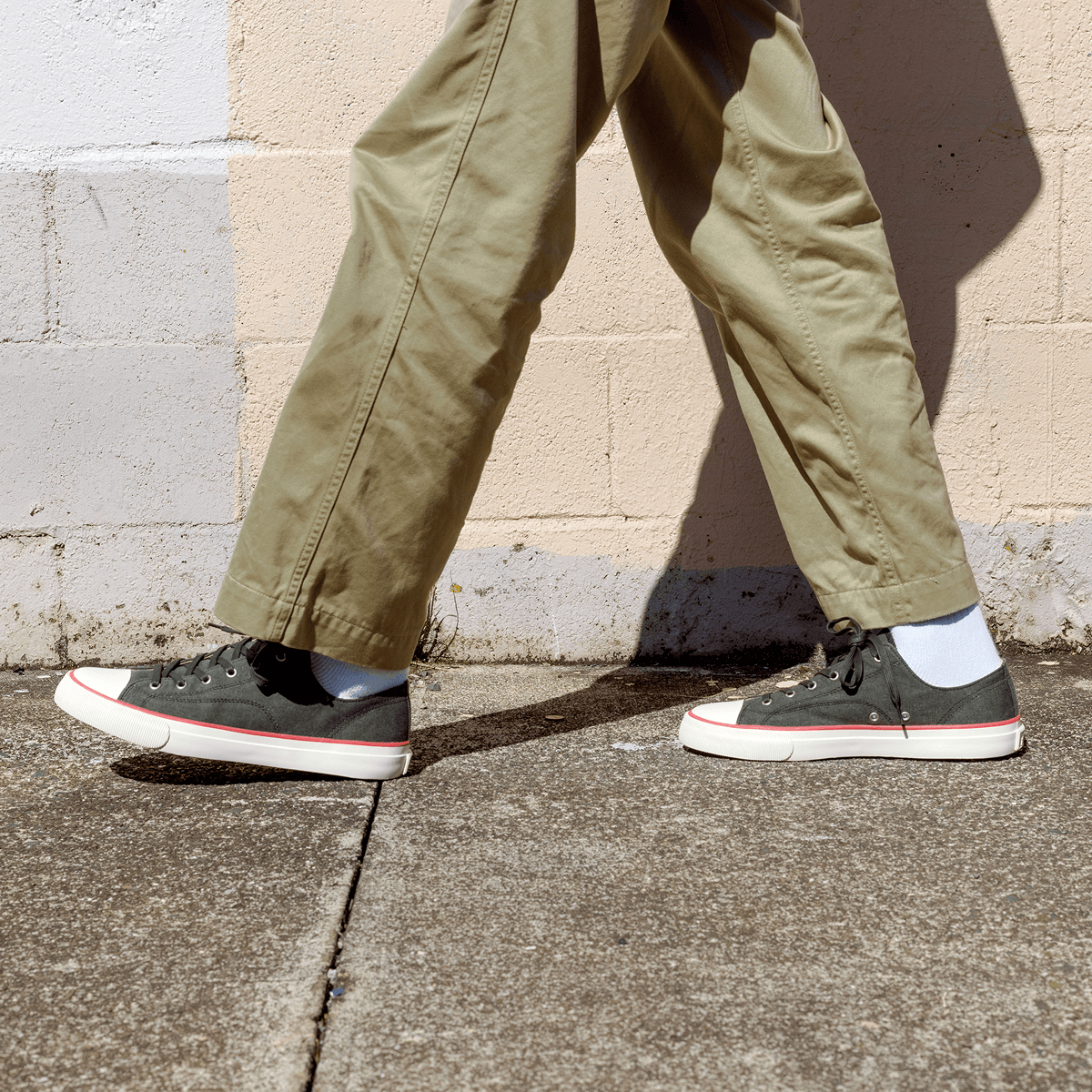 Person wearing black low-top sneakers and khaki pants against a stone wall.