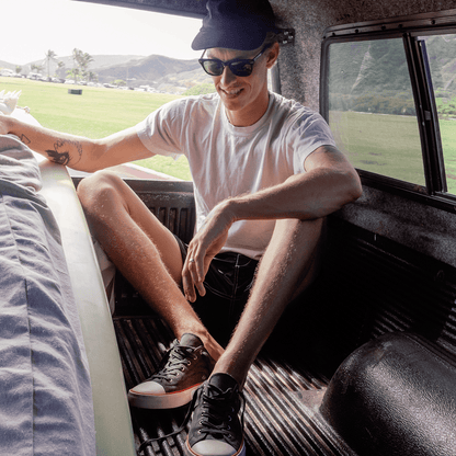 Man wearing sunglasses, black low-top sneakers and shorts sitting in the back of an SUV.