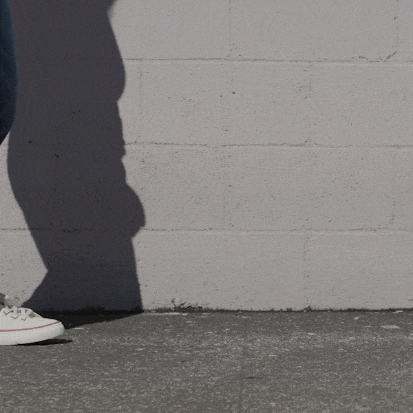 Person wearing white low-top sneakers and blue jeans shifting stance against a stone wall.