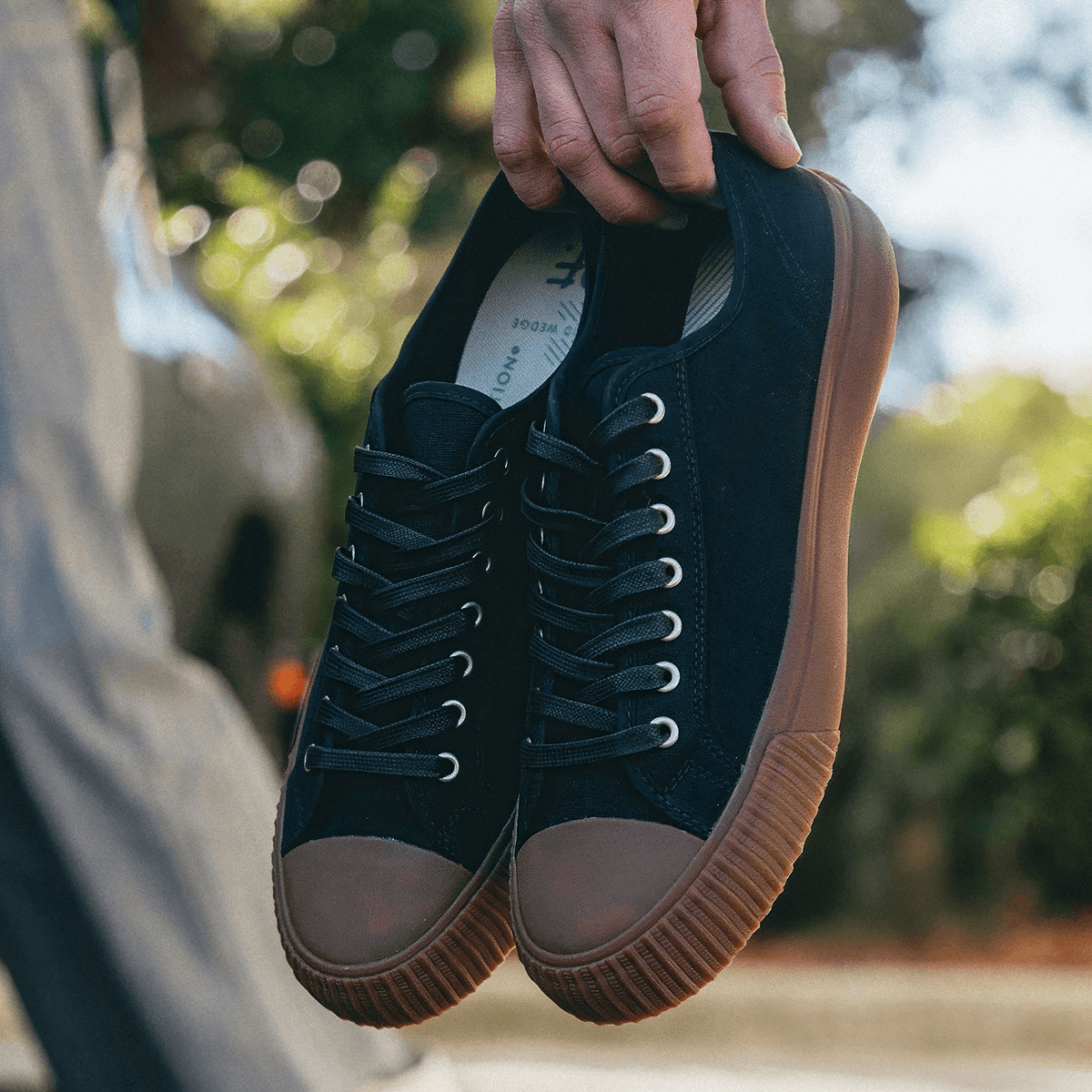 Person holding a pair of black low-top sneakers with brown soles outdoors.