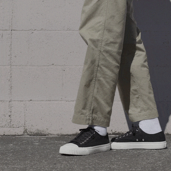 Person wearing black low-top sneakers and khaki pants shifting stance against a stone wall.