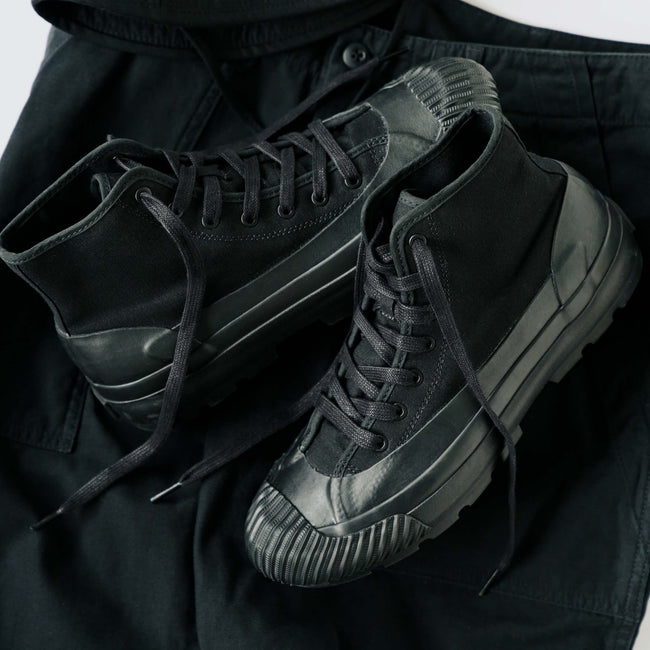 A pair of black boots with lugged rubber outsoles, displayed on top of a black pants against a light gray background.