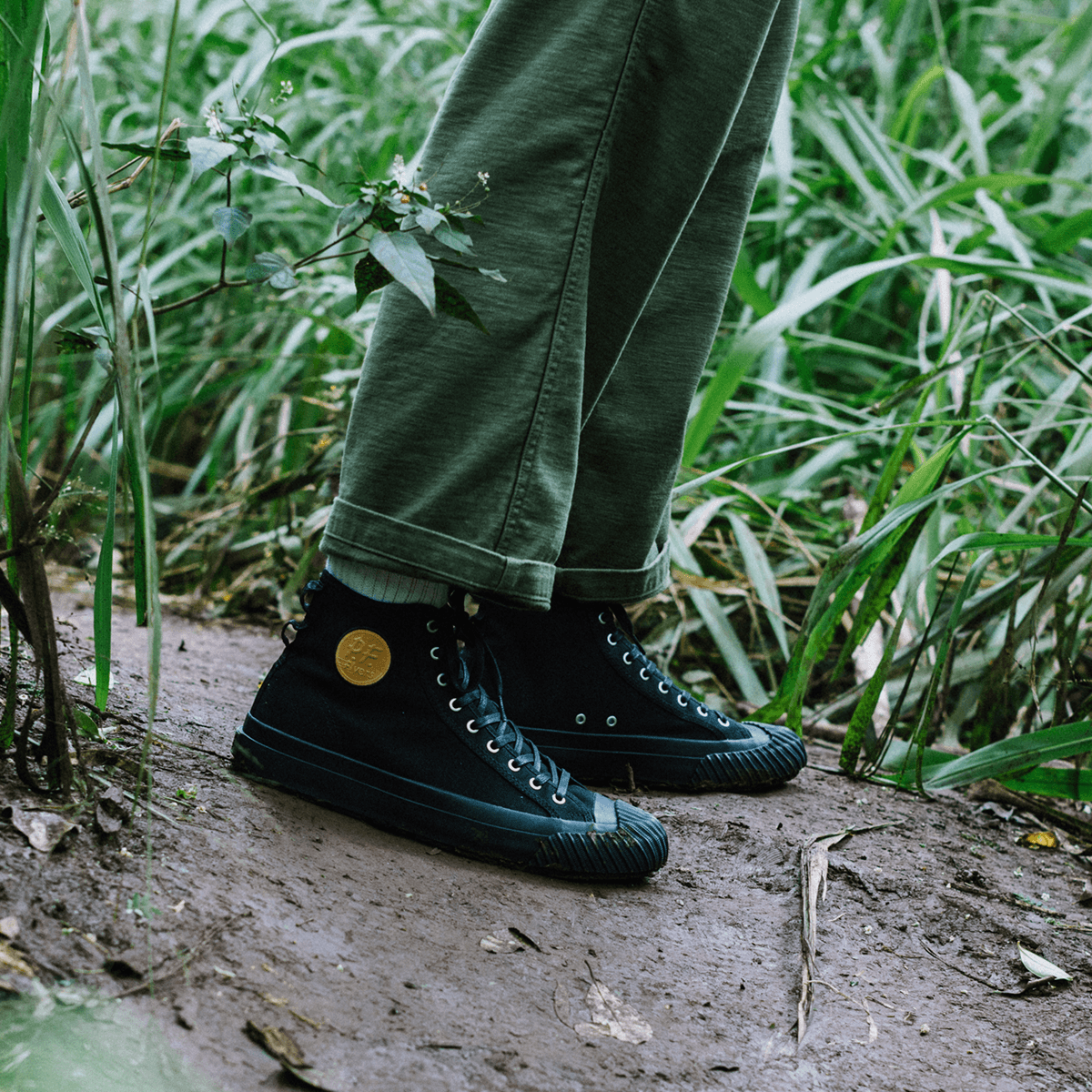 Person wearing black high-top sneakers standing on a muddy path surrounded by tall grass.
