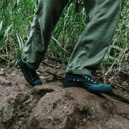 Person wearing green pants and black shoes walking on a muddy path.
