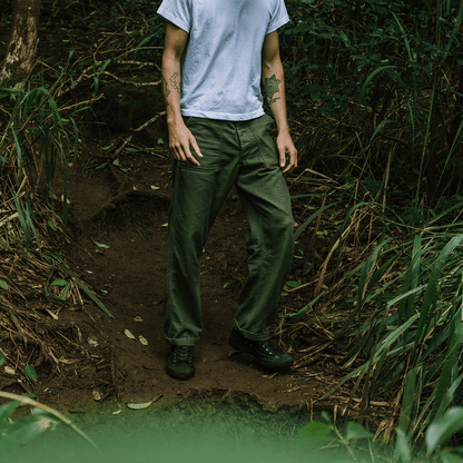 Man wearing a white t-shirt, black sneakers, and green pants standing in a forest.