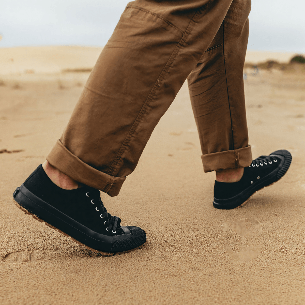 Person wearing black low-top sneakers and brown pants walking on a sand dune.