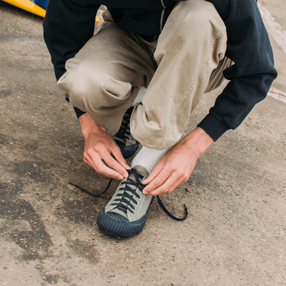 Person tying the black laces of an olive green sneaker outdoors.