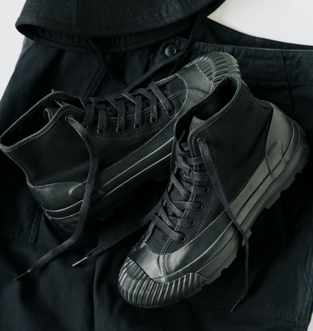 A pair of black boots with lugged rubber outsoles, displayed on top of a black pants against a light gray background.