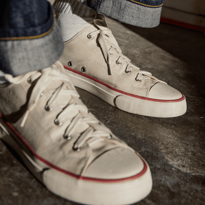 Close-up of white low-top sneakers with crimson accents worn by a person on a concrete floor.