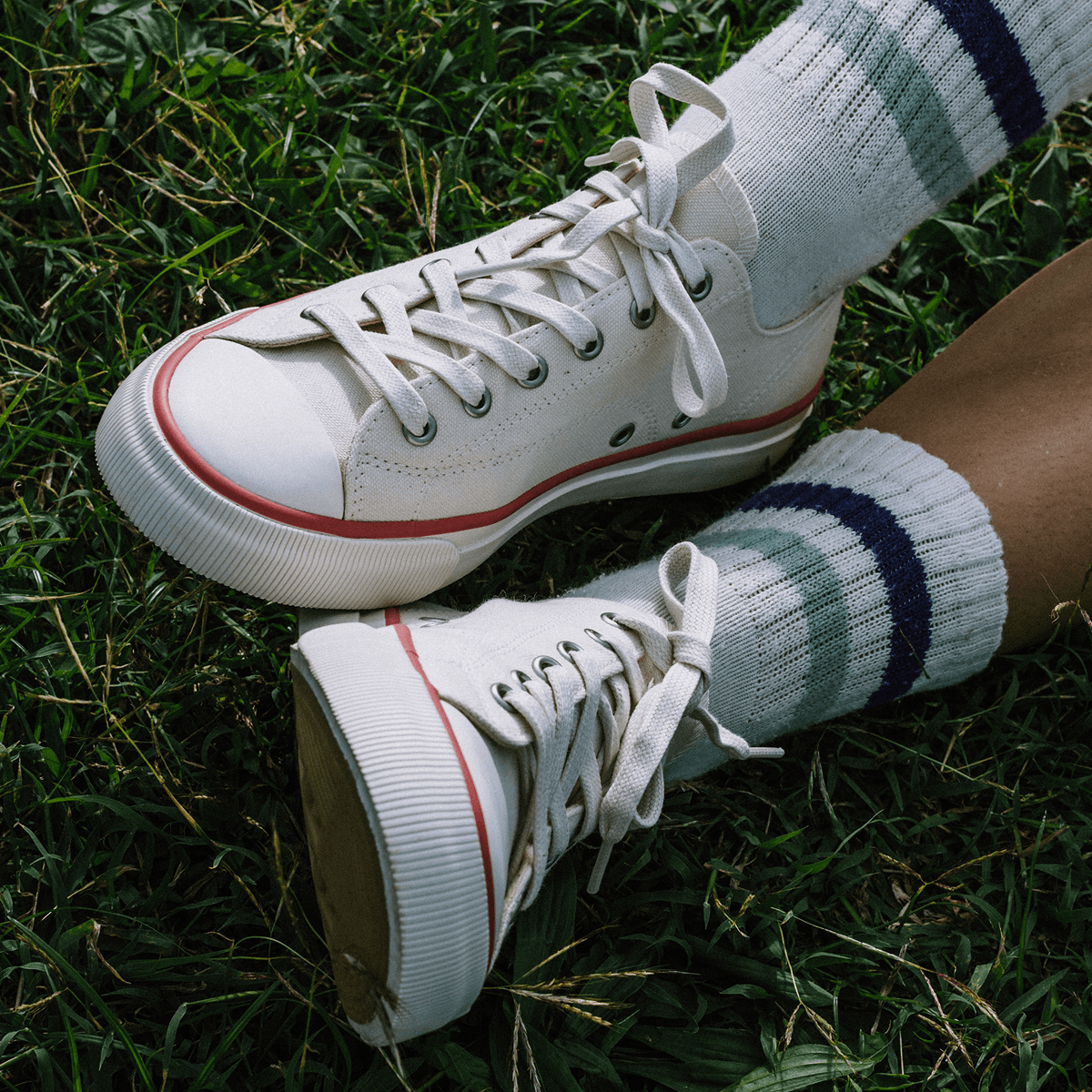 Close-up of white low-top sneakers with crimson accents, worn with white ankle socks on grass.