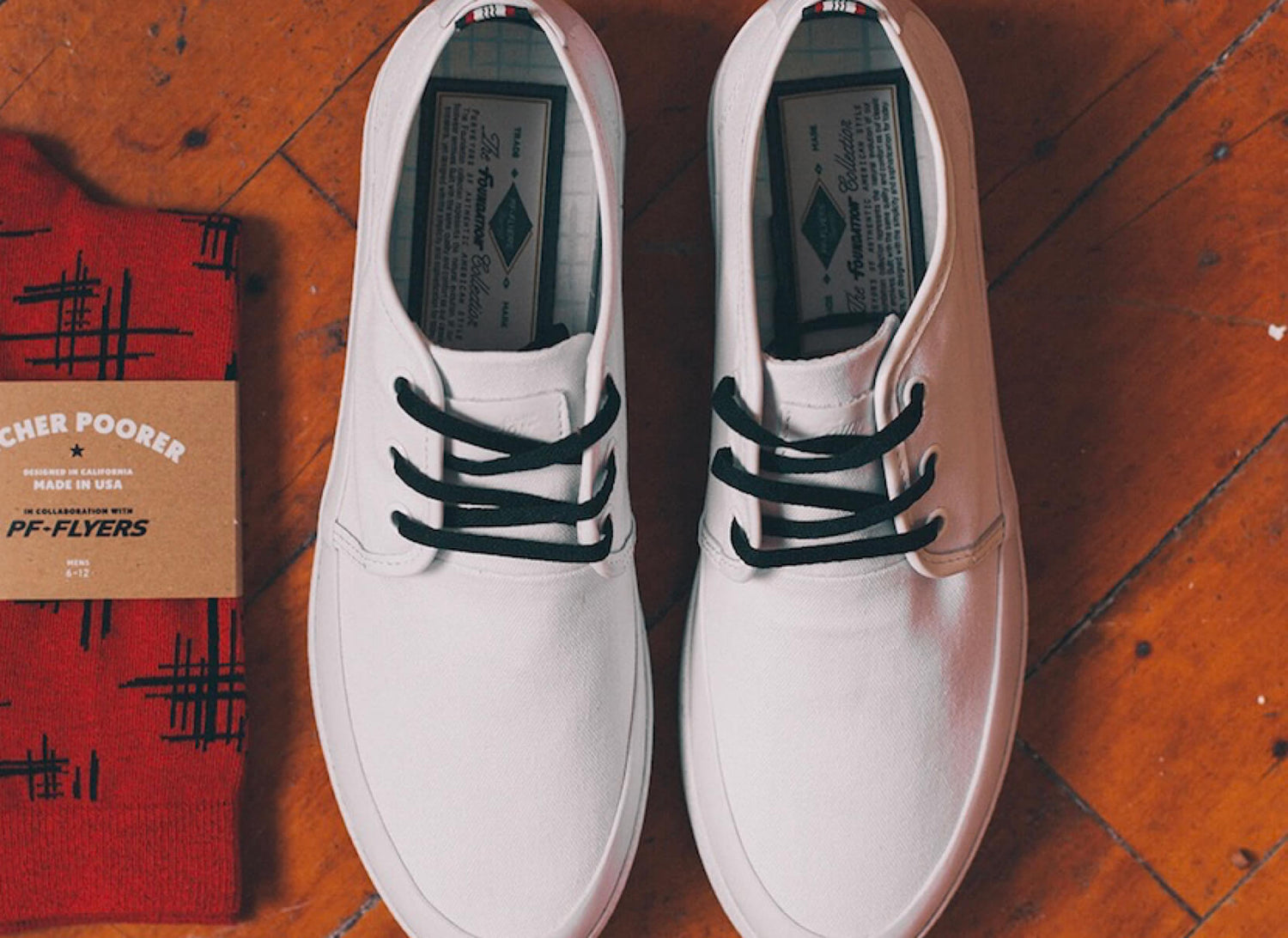 White retro low-top sneakers with black laces on a wooden floor beside a red P.F. Flyers sock with black patterns.