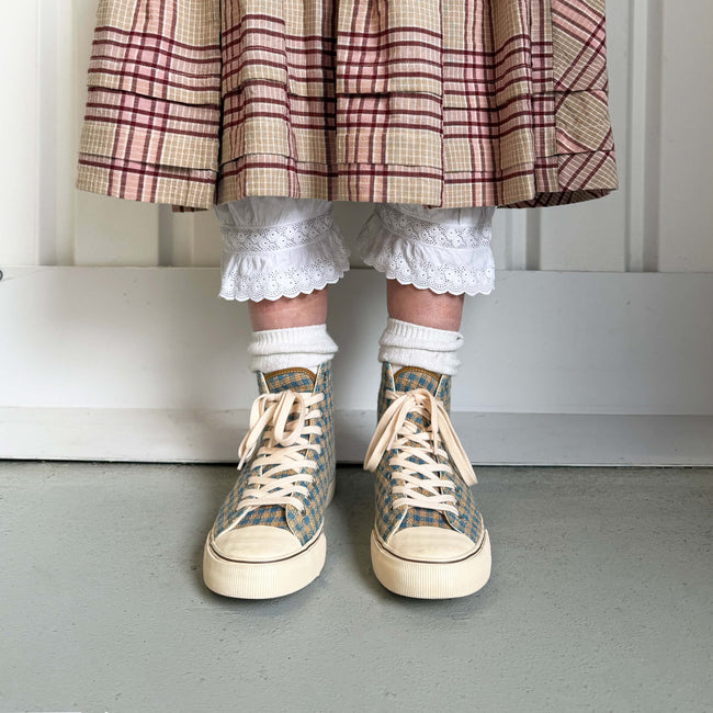 A person standing against a white paneled wall wearing indigo and brown plaid high-top sneakers.